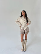 A woman modeling a cream and beige vertically striped button-up shirt, matching beige shorts with lace trim, and beige knee-high boots against a plain light gray studio background.