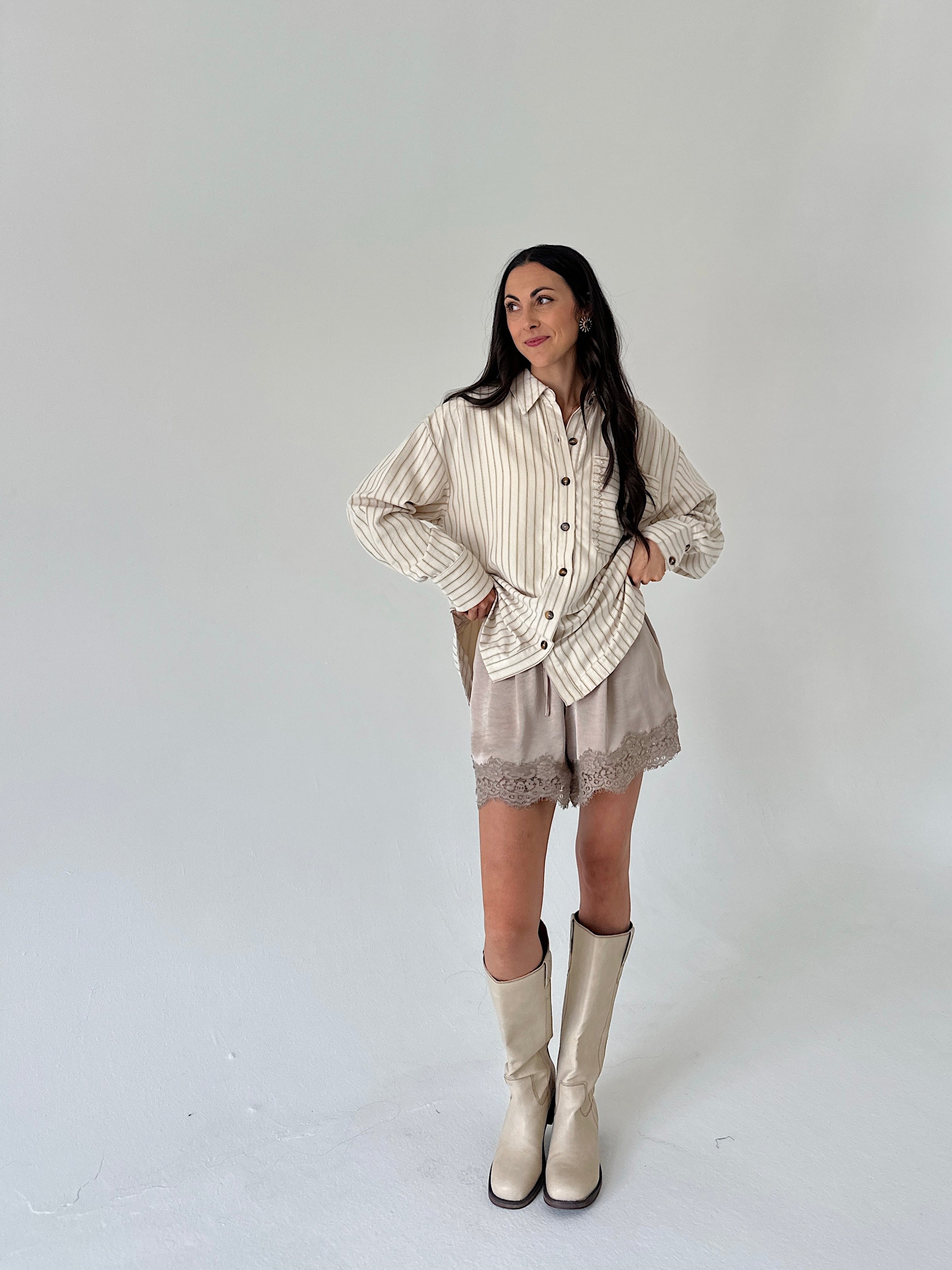 A woman modeling a cream and beige vertically striped button-up shirt, matching beige shorts with lace trim, and beige knee-high boots against a plain light gray studio background.