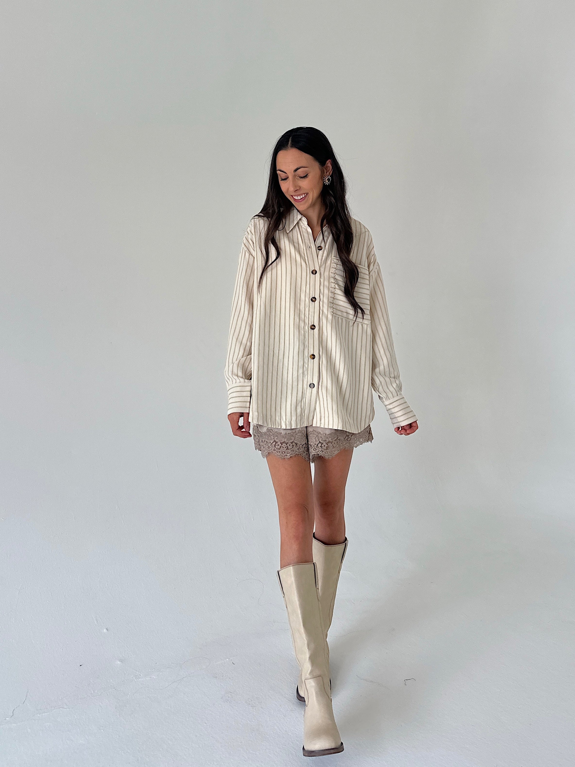 A woman modeling a cream and beige vertically striped button-up shirt with lace shorts and beige knee-high boots, standing against a plain light gray studio background.