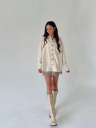 A woman modeling a cream and beige vertically striped button-up shirt with lace shorts and beige knee-high boots, standing against a plain light gray studio background.