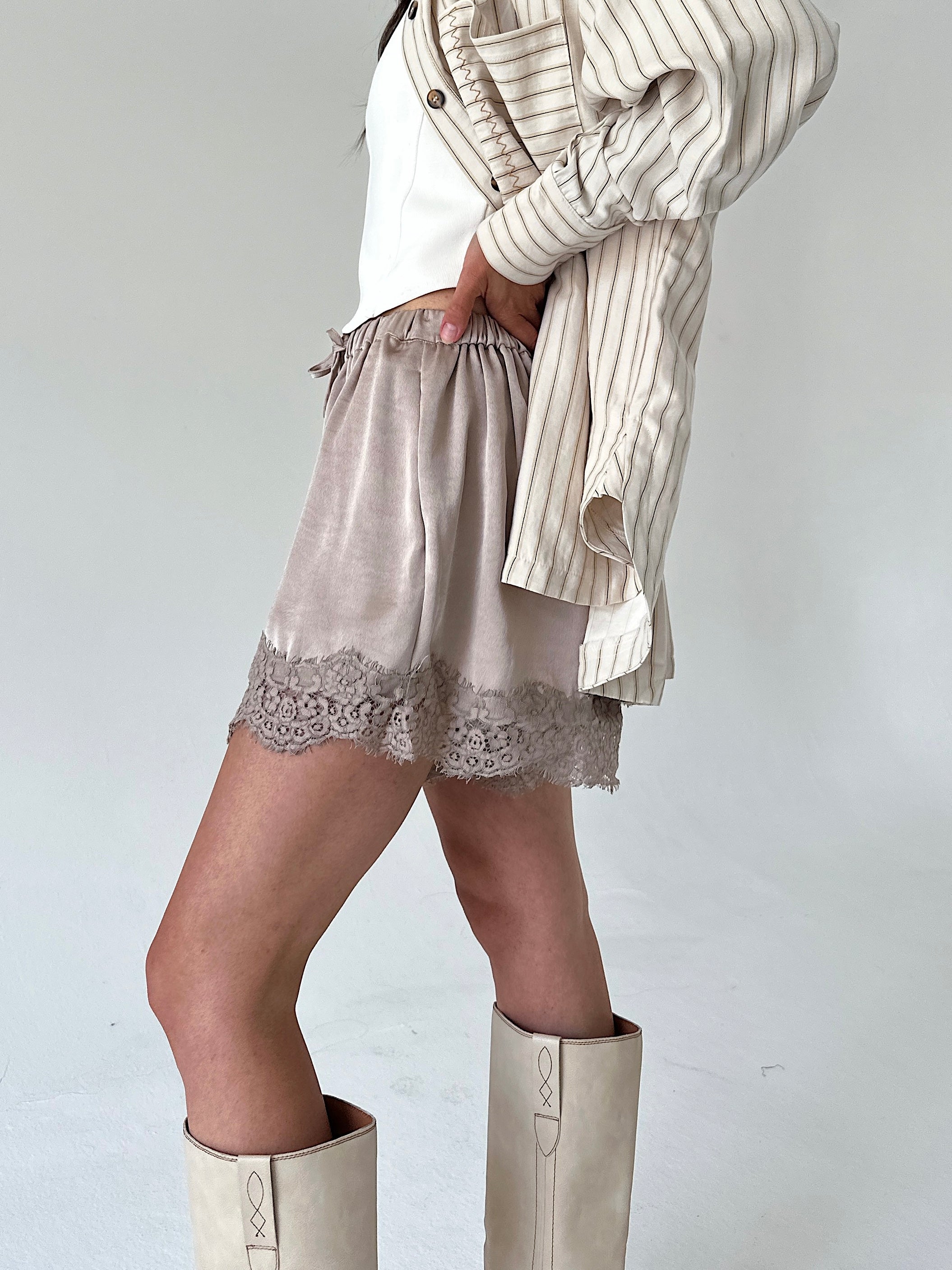 A model wearing beige satin shorts with a lace hem, styled with a white top, a striped button-up shirt draped over the arm, and beige boots against a plain light gray background.