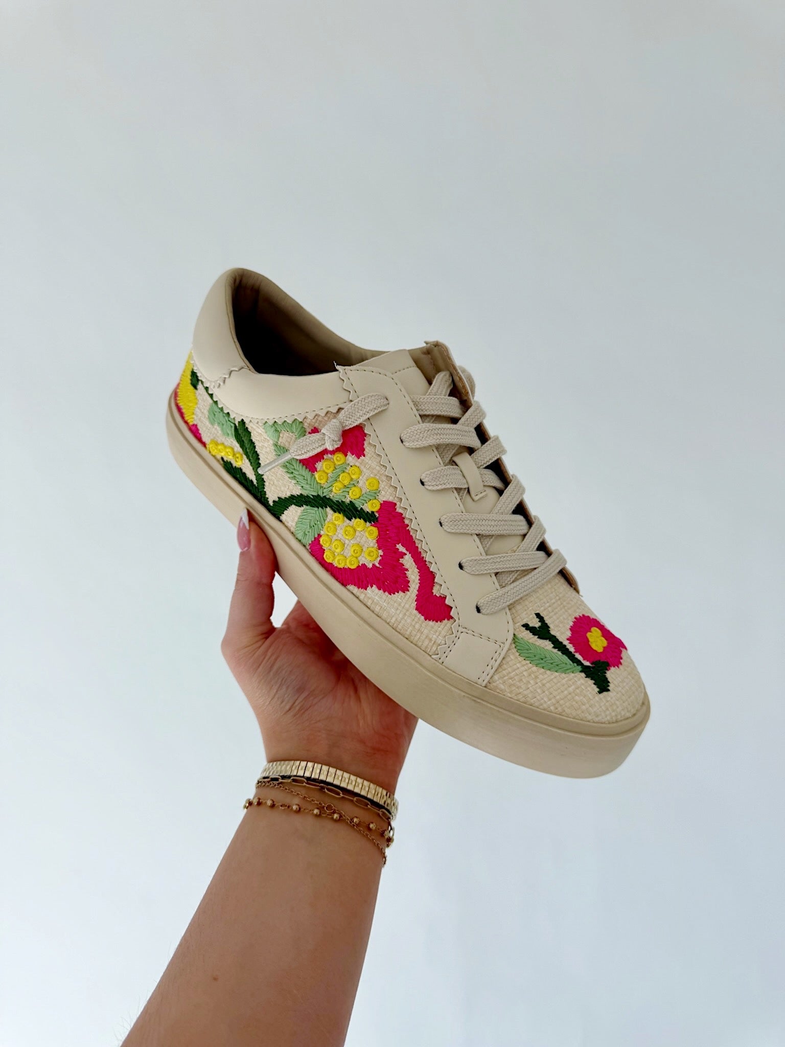 Beige sneaker with floral embroidery held by a hand against a light background