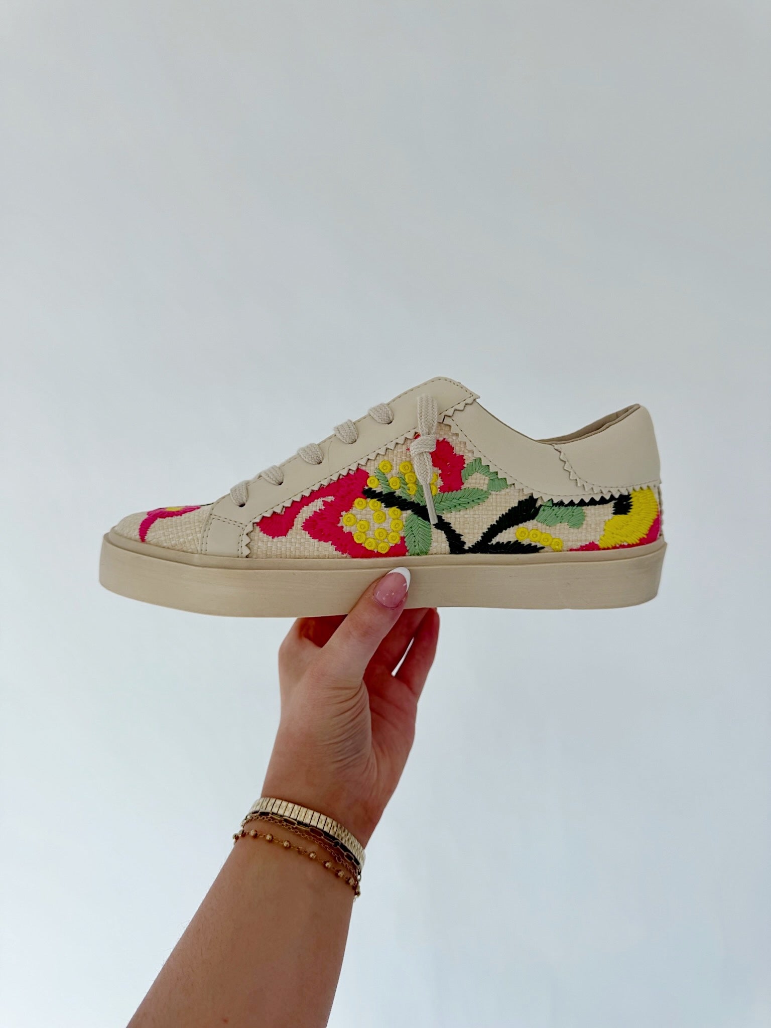 Beige sneaker with colorful floral embroidery held by a hand against a light gray background
