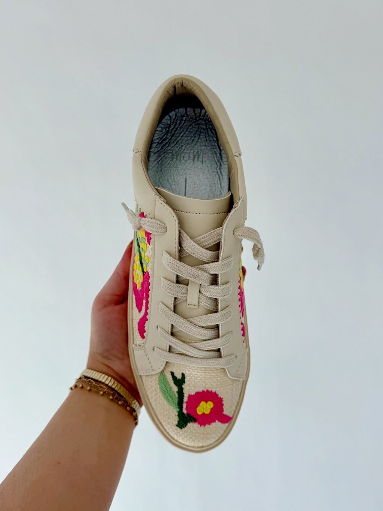 Beige sneaker with floral embroidery held by a hand against a light gray background