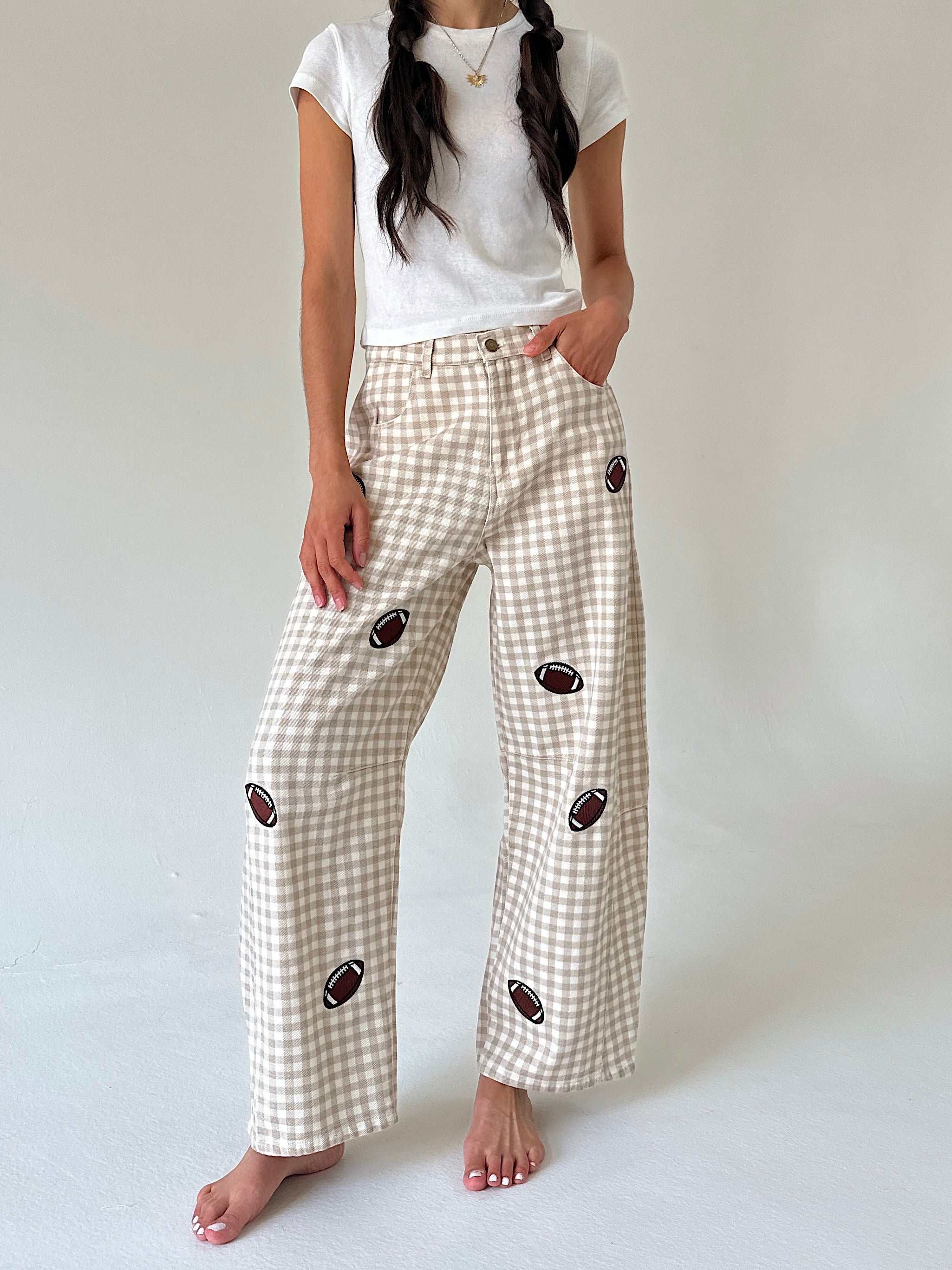 A model wearing beige and white gingham wide-leg pants with embroidered football motifs, paired with a white t-shirt, standing against a neutral light gray background.
