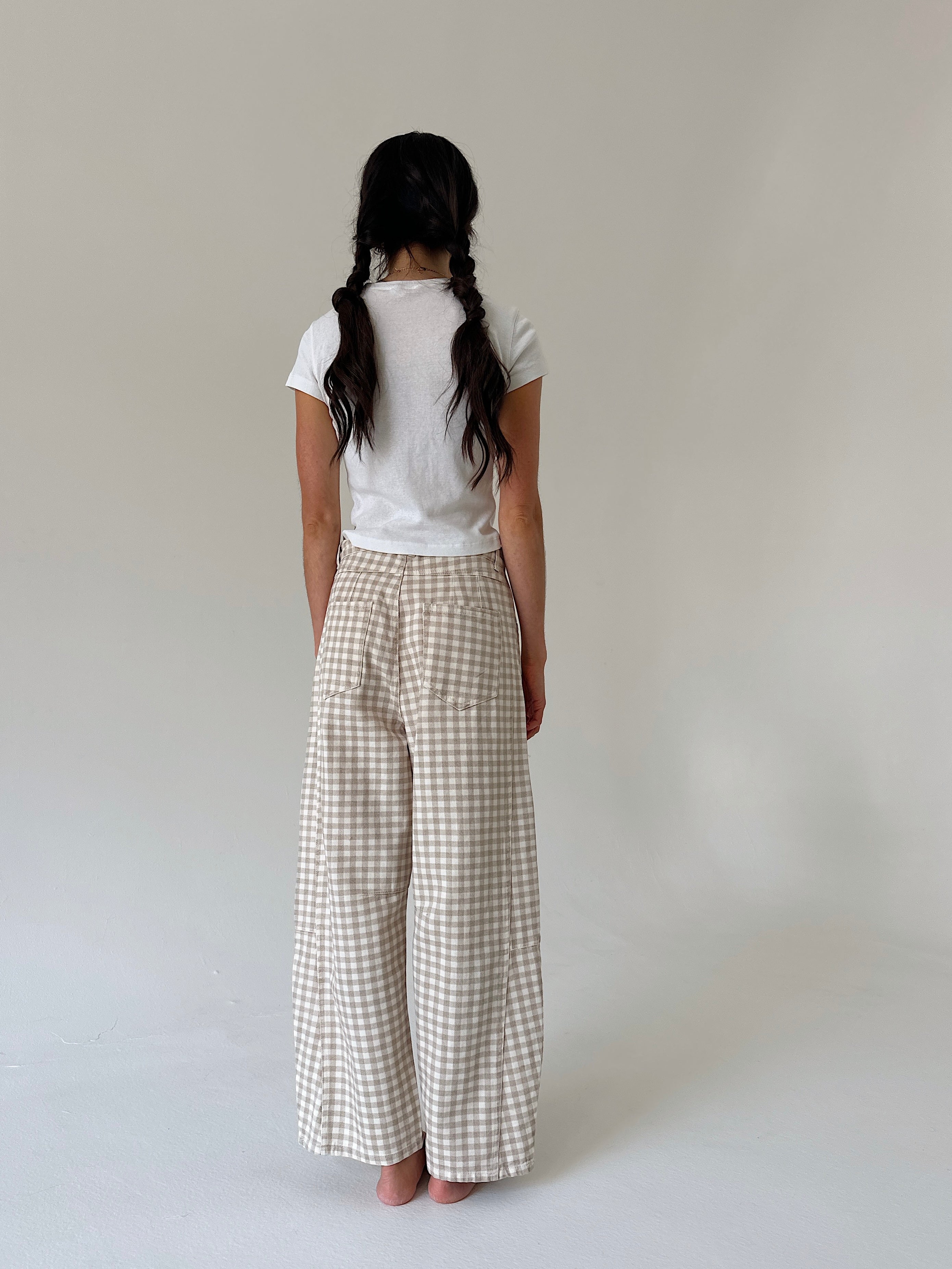 A woman viewed from behind wearing a white t-shirt and beige and white gingham wide-leg trousers, standing against a plain light gray studio background.