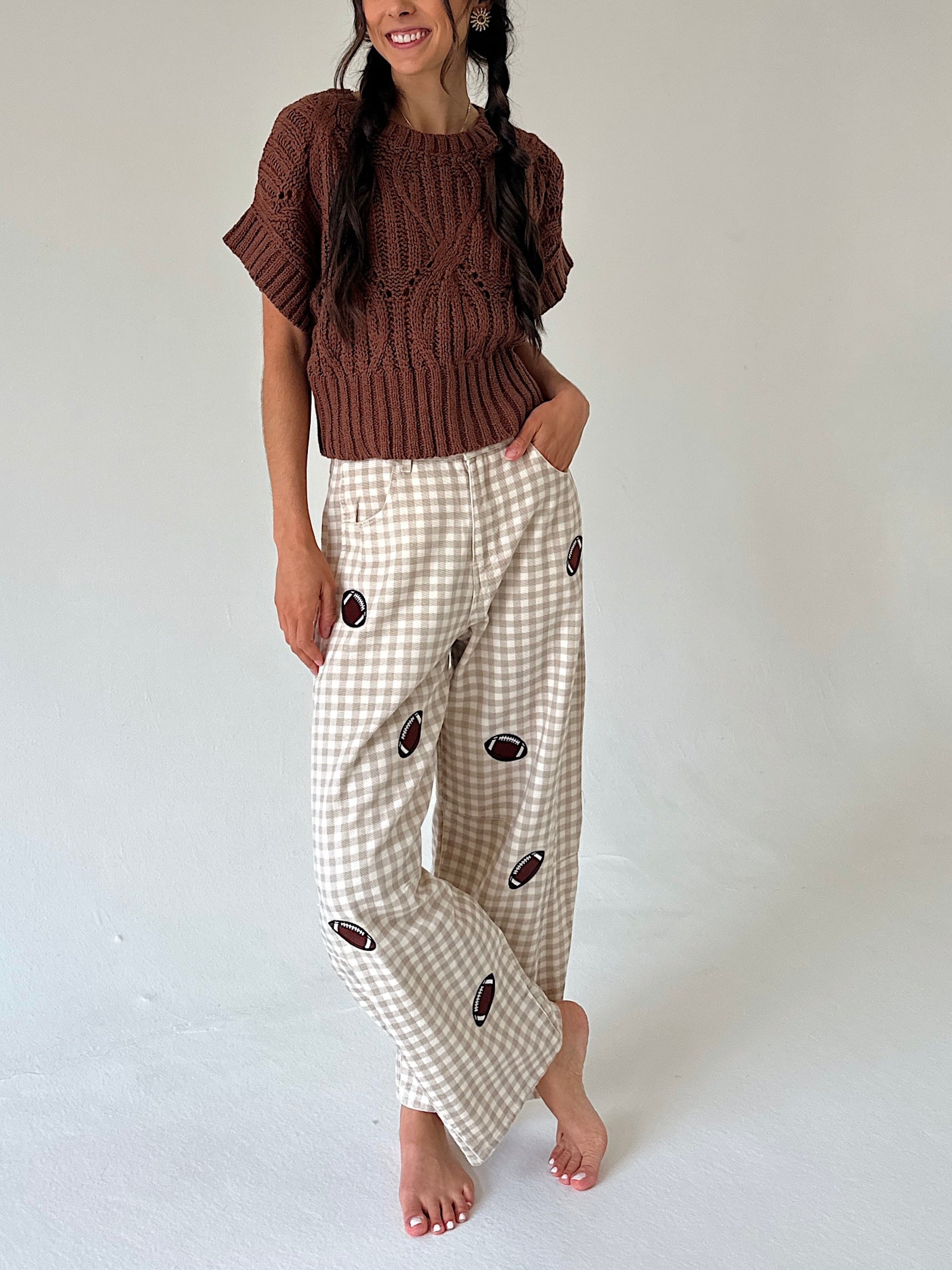 A woman modeling a brown short-sleeve knit sweater and beige gingham pants with football prints, standing against a plain light gray studio background.
