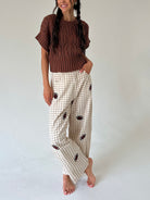 A woman modeling a brown short-sleeve knit sweater and beige gingham pants with football prints, standing against a plain light gray studio background.