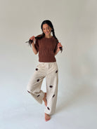 A woman posing in a studio setting, wearing a brown cable-knit short-sleeve top and cream gingham wide-leg pants with dark brown embroidered details.