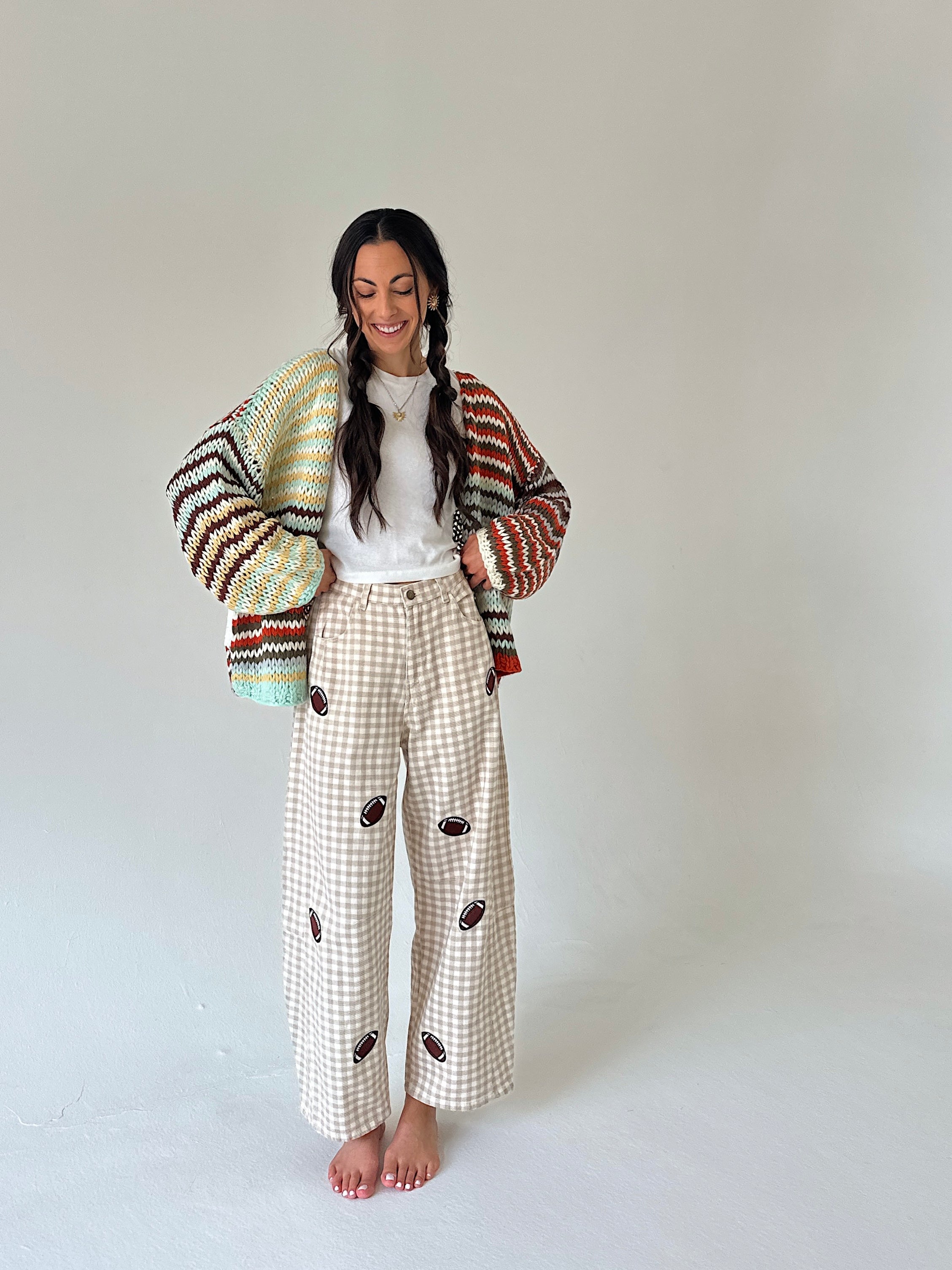 A woman stands barefoot against a plain light gray studio background, wearing a multicolor knit cardigan draped off her shoulders, a white top, and cream gingham wide-leg pants with brown embroidered details.