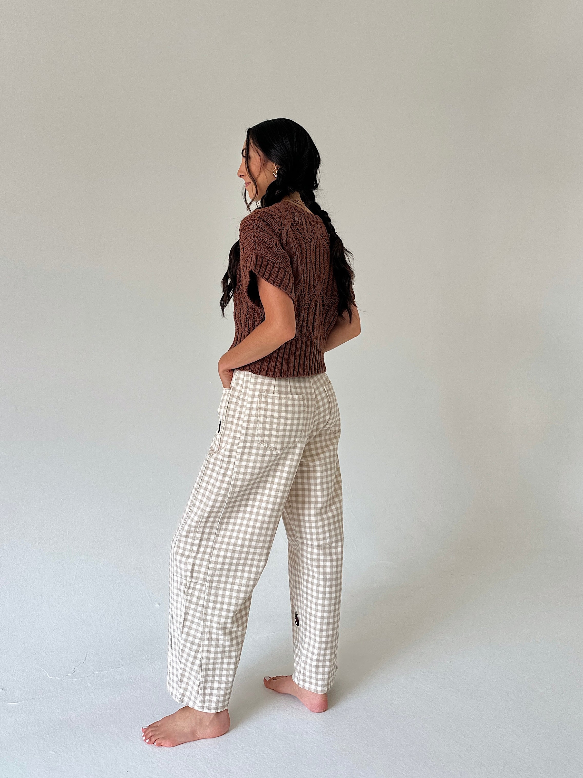 A woman stands barefoot in profile wearing a brown short-sleeve knit top and cream-and-white gingham wide-leg pants against a plain light gray studio background.