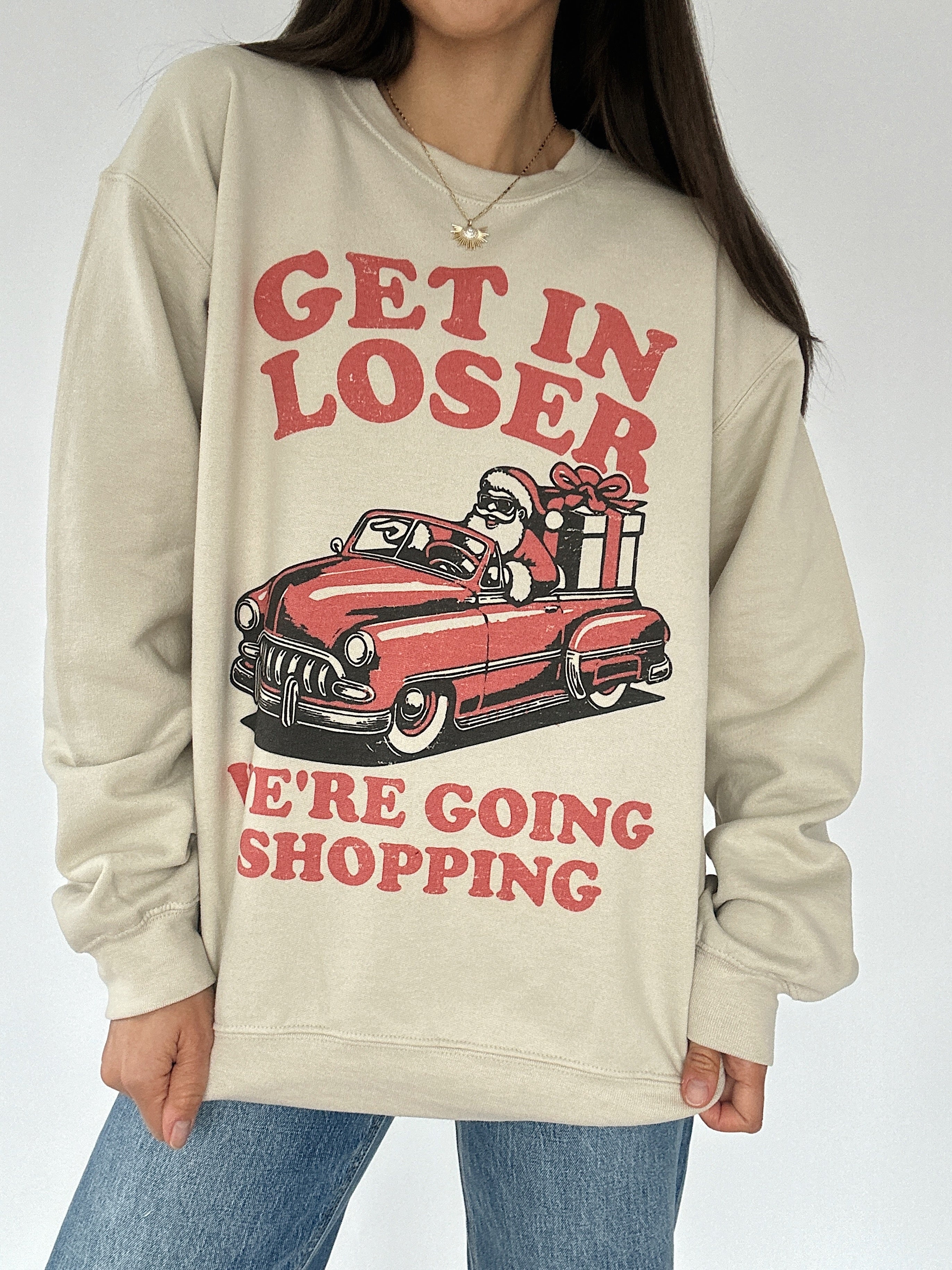 Woman wearing a sand sweatshirt with a car graphic that has Santa inside with words surrounding that says "Get in loser we're going shopping"