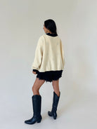 A woman is shown from behind wearing a cream-colored oversized sweater, a black fringed mini skirt, and black leather cowboy boots, set against a plain off-white studio background.