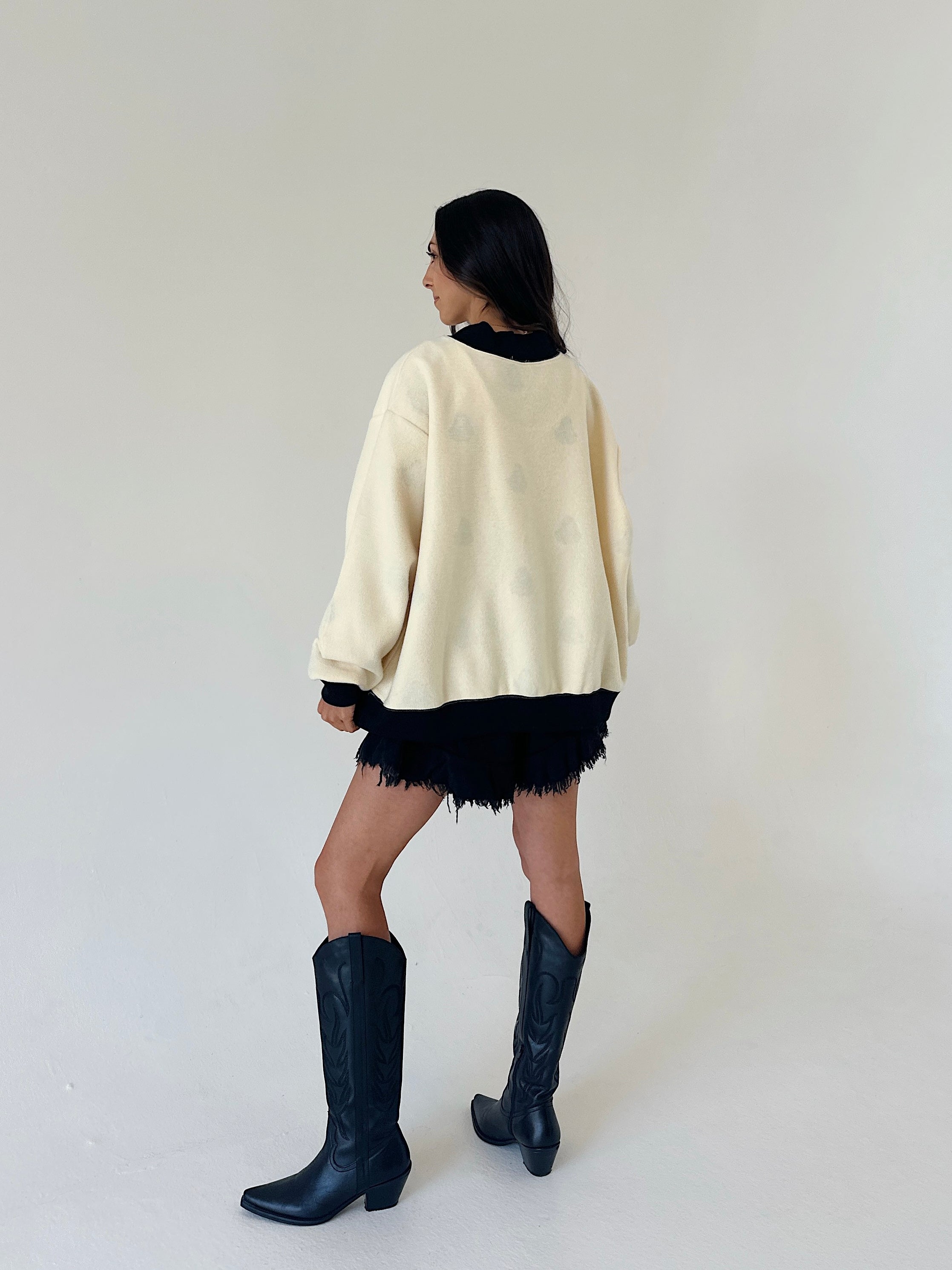 A woman is shown from behind wearing a cream-colored oversized sweater, a black fringed mini skirt, and black leather cowboy boots, set against a plain off-white studio background.