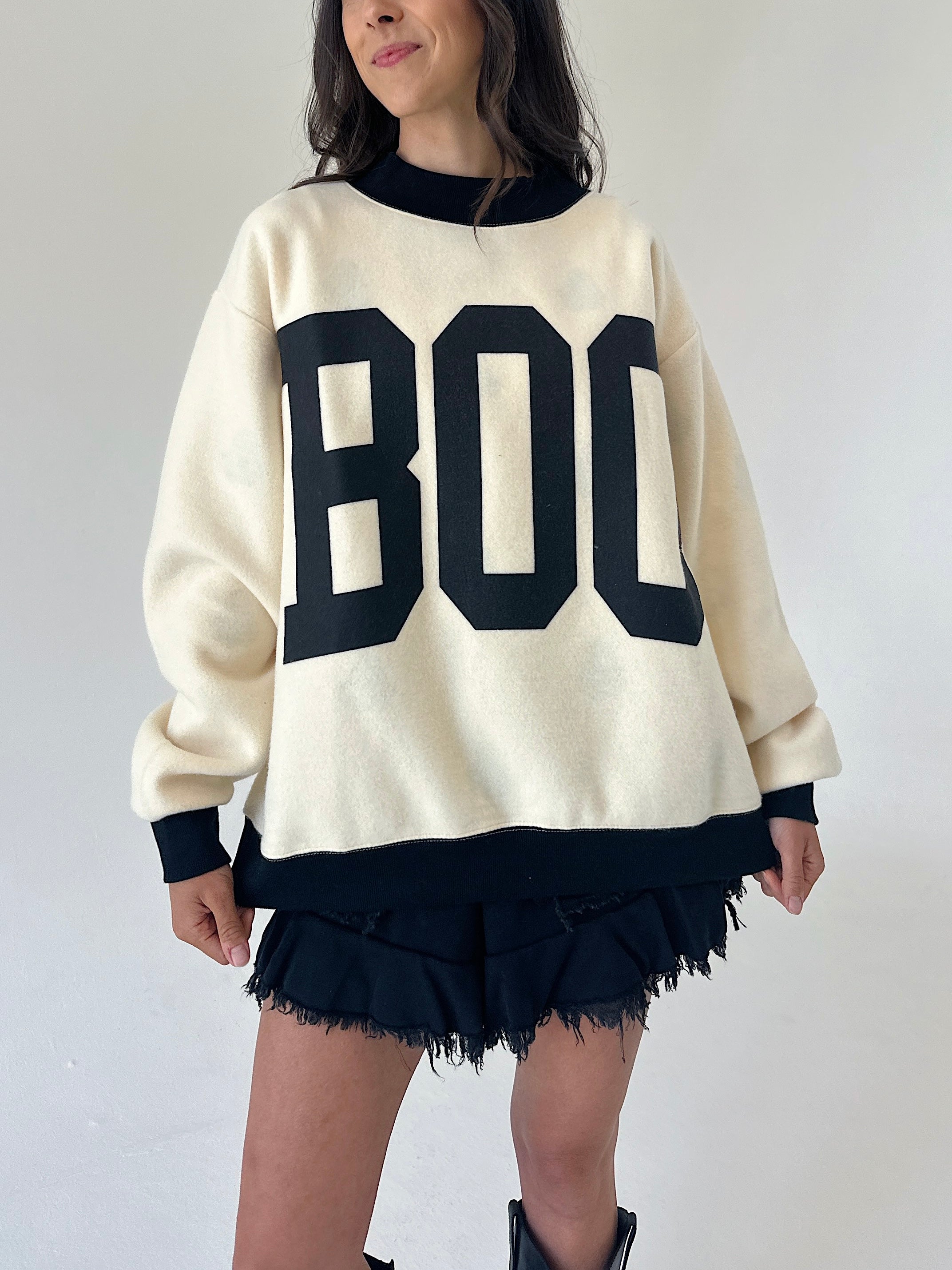 A woman wearing an oversized cream-colored sweater with large black block letters spelling 'BOO' across the front, paired with black frayed shorts and black boots, set against a plain light gray wall.