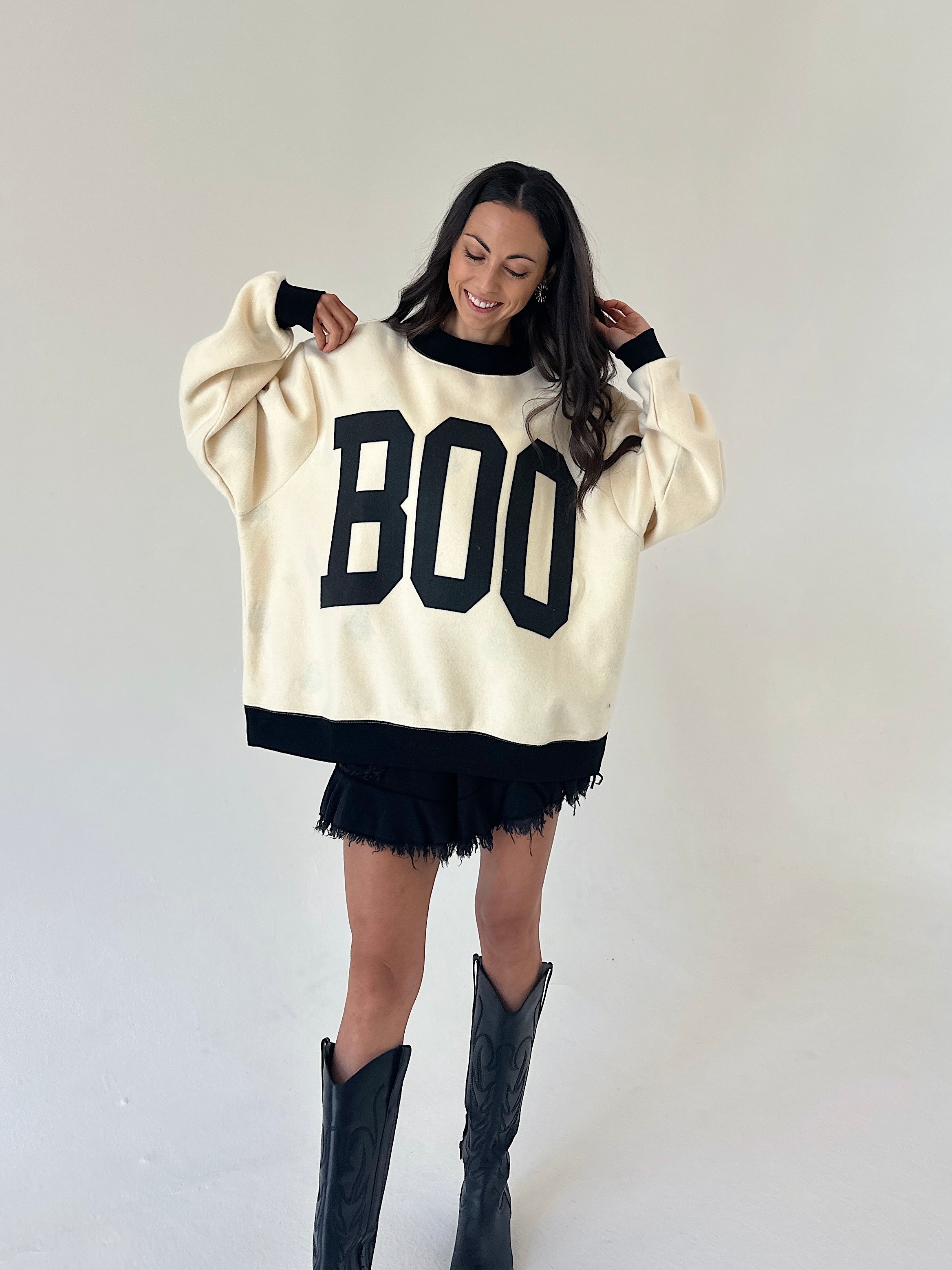 A woman modeling a cream-colored knit sweater with black trim and large black 'BOO' lettering on the front, paired with black shorts and black cowboy boots, against a plain light gray studio background.