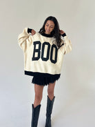 A woman modeling a cream-colored knit sweater with black trim and large black 'BOO' lettering on the front, paired with black shorts and black cowboy boots, against a plain light gray studio background.