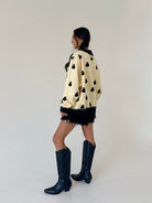 woman standing from a back side angle wearing a cream sweatshirt covered in black ghost graphics, wearing black shorts and tall black boots