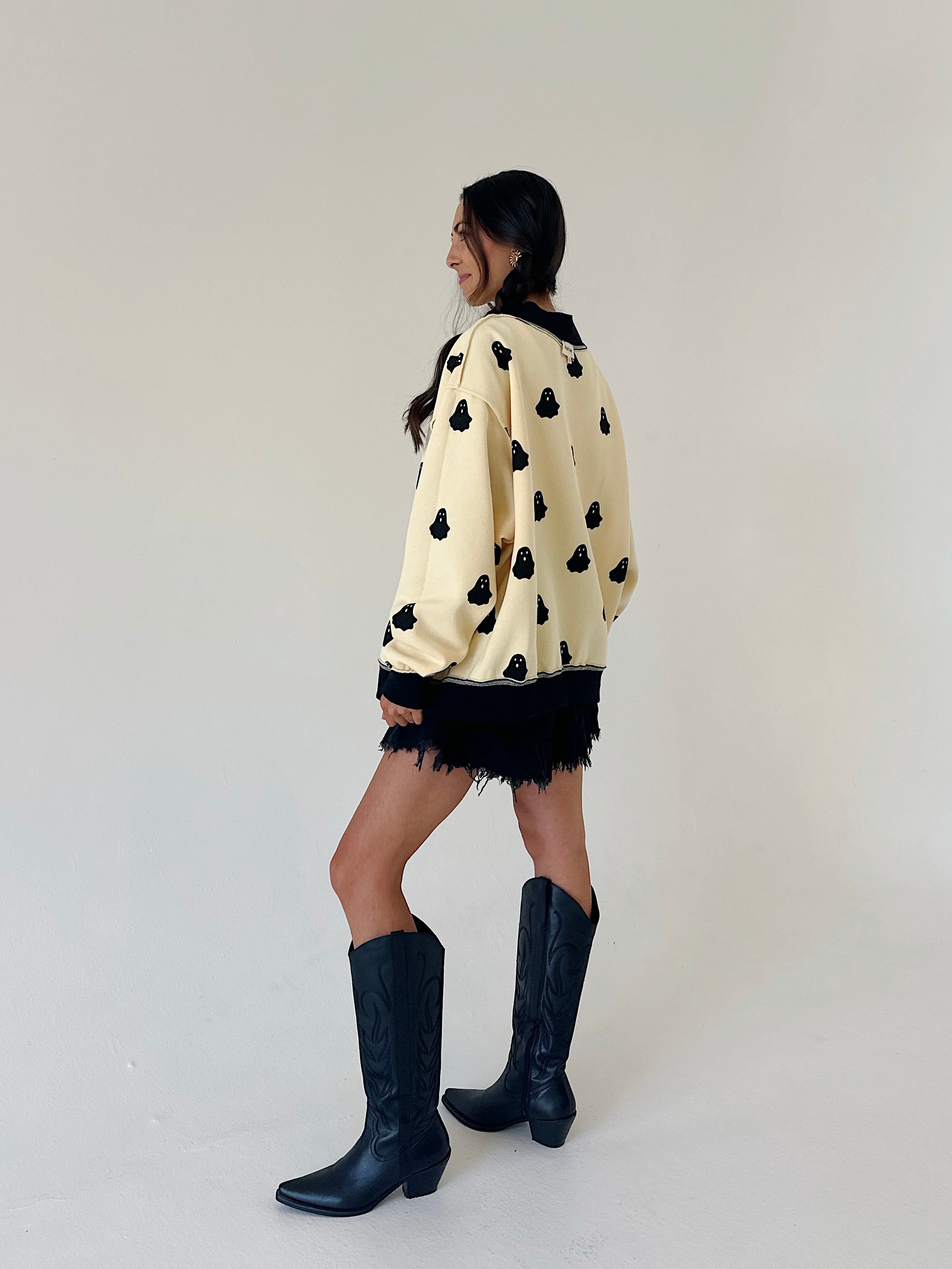 woman standing from a back side angle wearing a cream sweatshirt covered in black ghost graphics, wearing black shorts and tall black boots