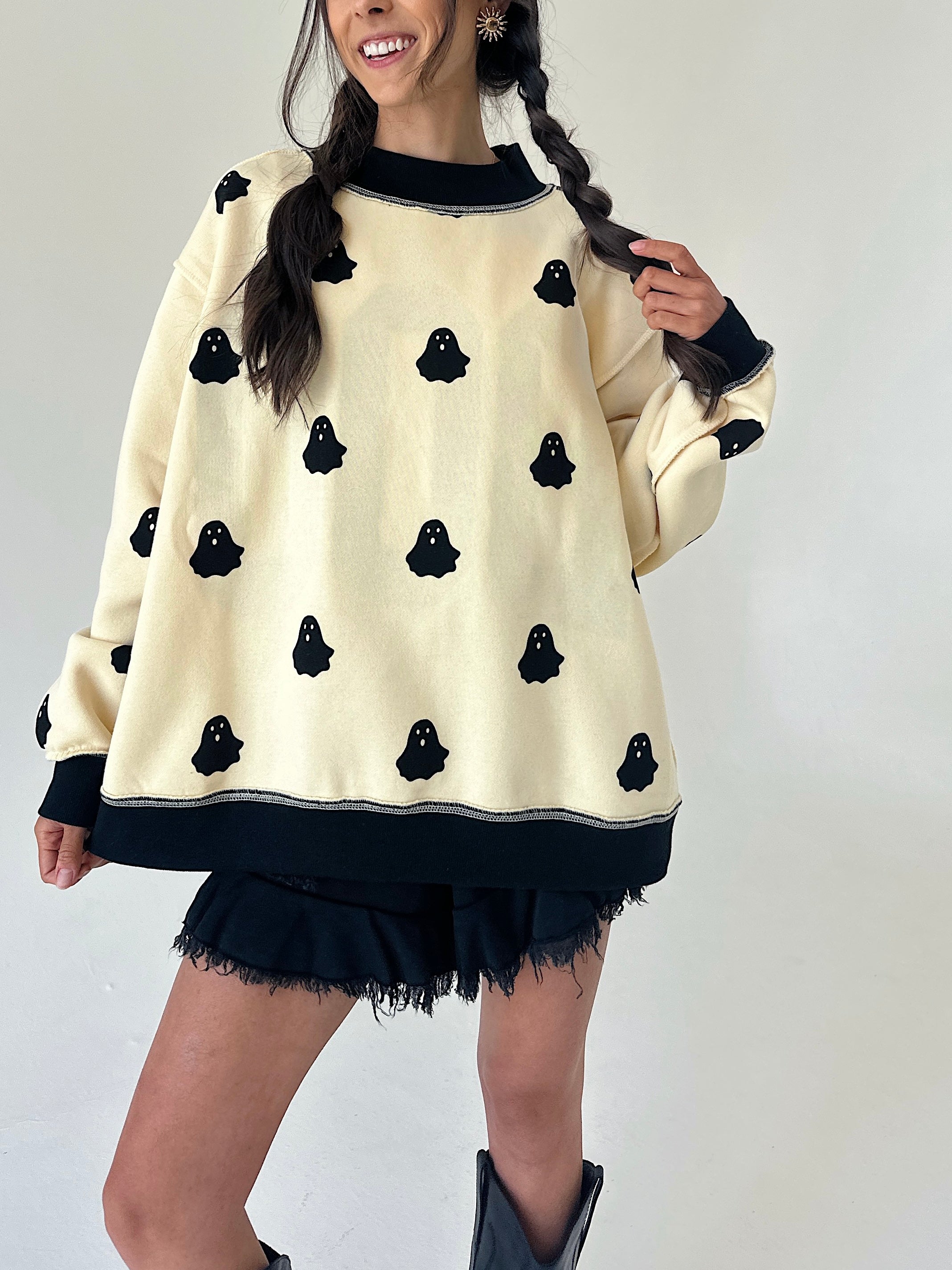 A woman wearing a cream-colored oversized sweater with black ghost motifs and black ribbed trim, paired with black frayed shorts and black boots, set against a plain light background.