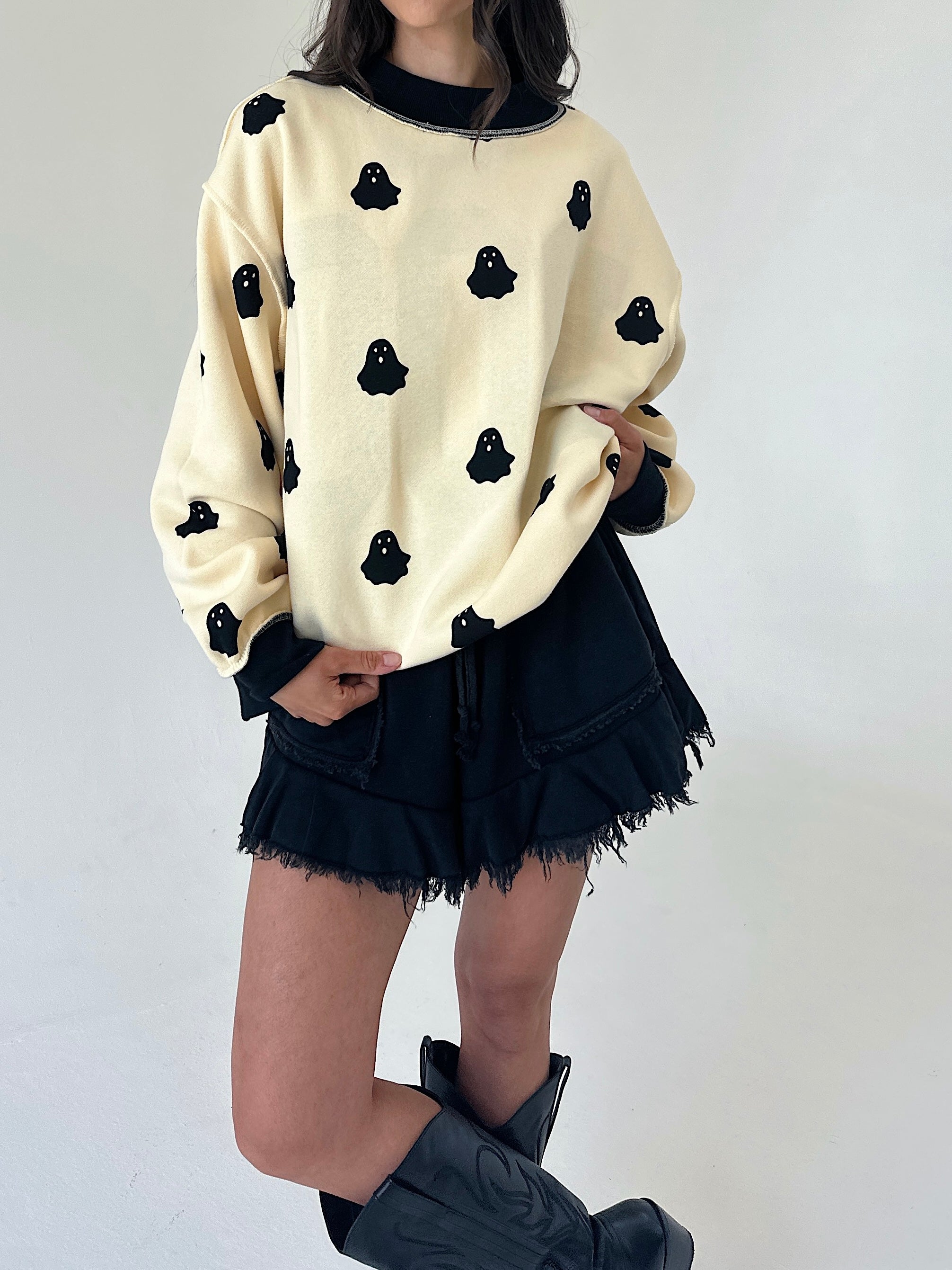 A person wearing a cream-colored crewneck sweater with black ghost motifs, paired with black frayed denim shorts and black boots, standing against a plain white wall.
