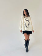 A woman modeling a cream-colored knit sweater with a black ghost graphic, paired with a black ruffled mini skirt and black cowboy boots, against a plain off-white studio background.