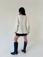 Woman standing from the back wearing a cream oversized sweater over black lace shorts with tall black boots