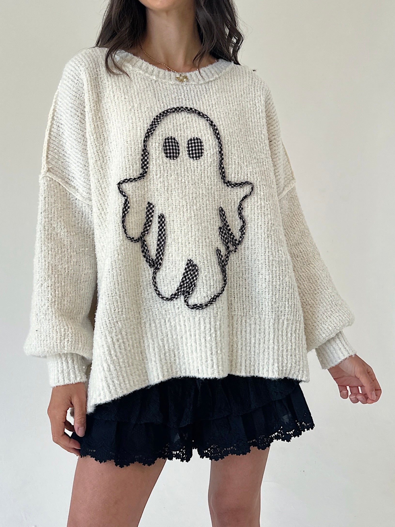 A person wearing an oversized cream knit sweater with a black ghost graphic on the front, paired with a black ruffled skirt, against a plain light background.