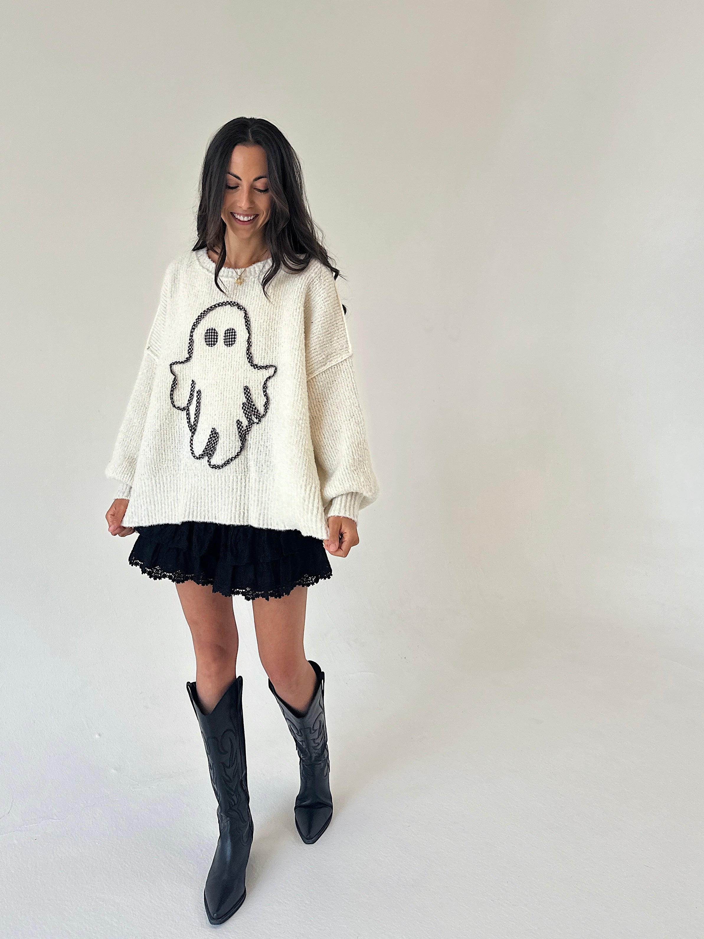 A woman modeling a cream-colored knit sweater with a black ghost graphic, paired with a black ruffled mini skirt and black cowboy boots, against a plain light gray studio background.