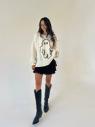 A woman modeling a cream knit sweater with a black ghost graphic, paired with a black ruffled mini skirt and black knee-high boots, standing against a plain off-white studio background.
