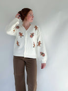 Person wearing a white cardigan with patch gingerbread men designs with brown pants on a plain background