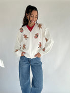 Woman wearing a white cardigan with gingerbread man patterns and blue jeans on a plain background