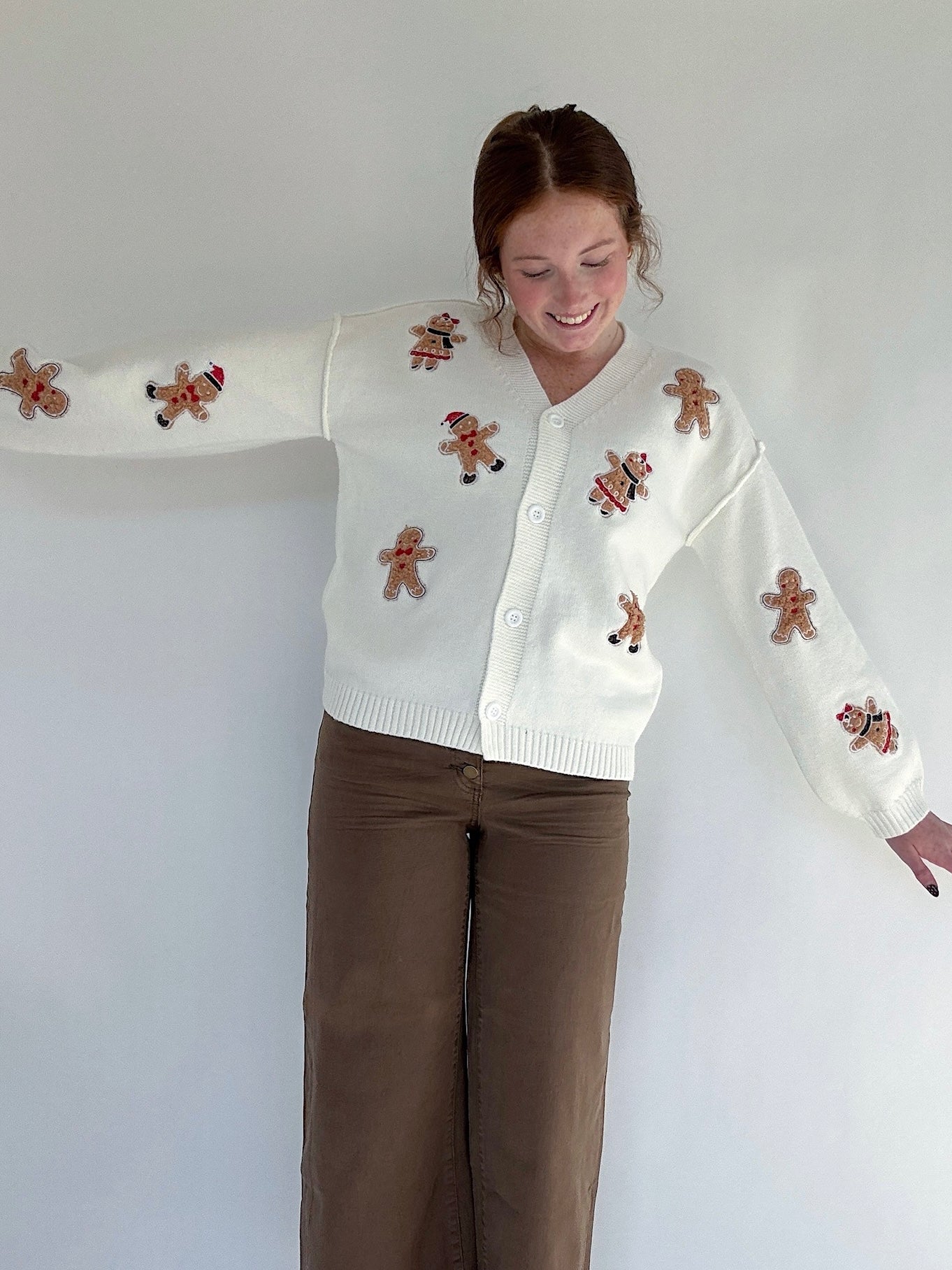 Person wearing a white sweater with gingerbread man patches and brown pants on a plain background
