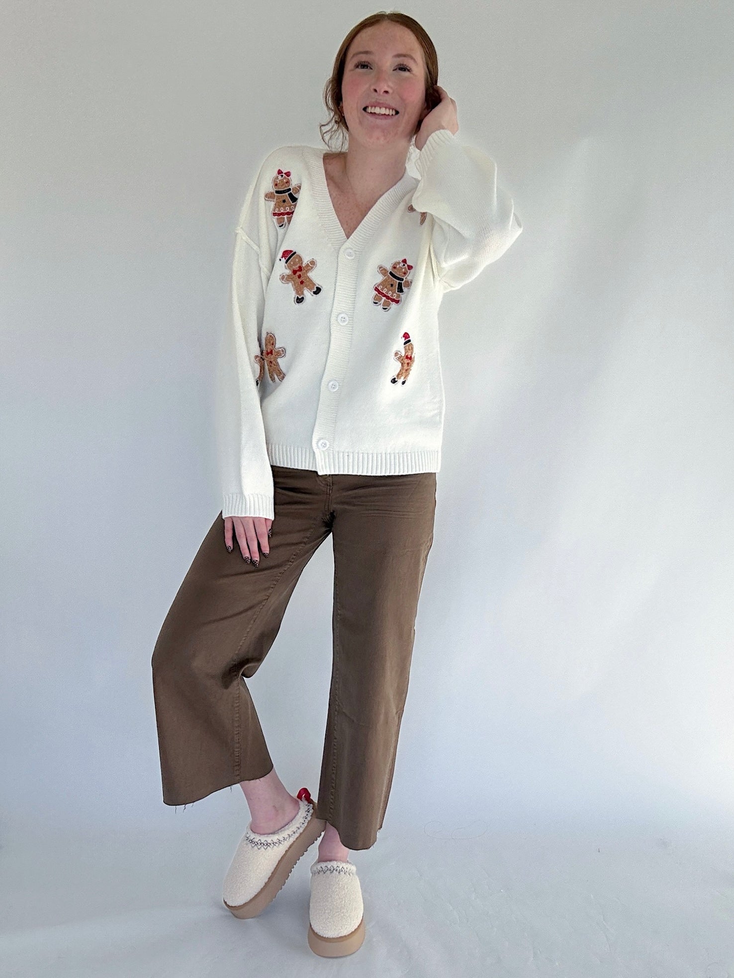 Person wearing a white cardigan with gingerbread man patterns and brown pants on a plain background