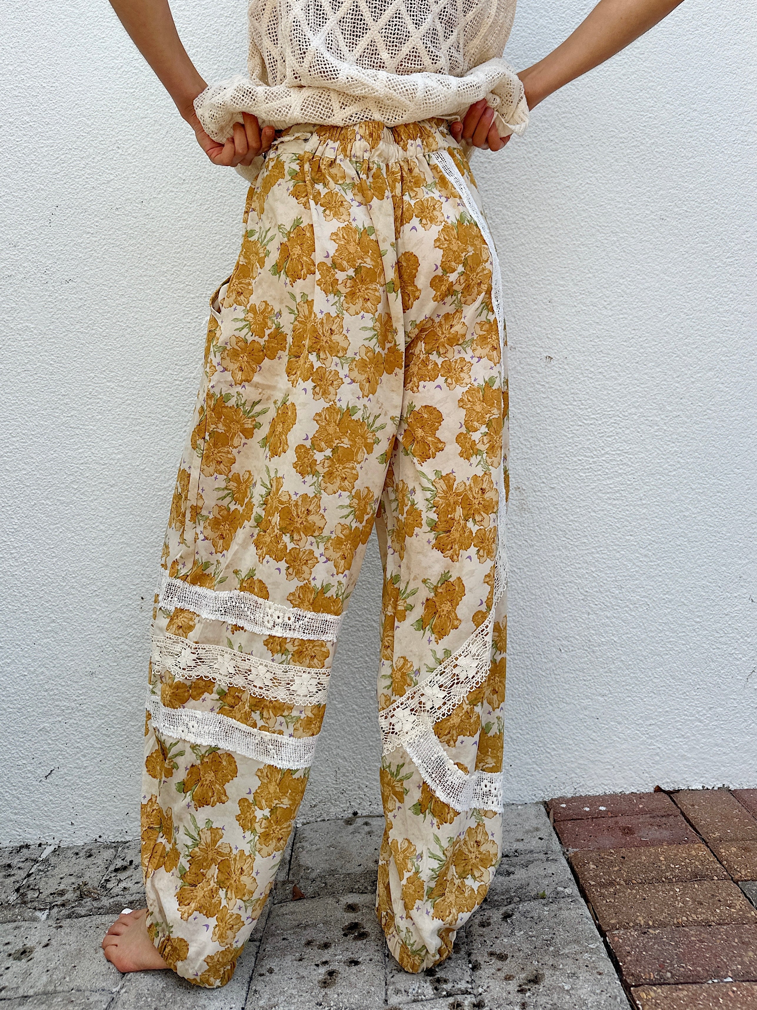 Person wearing mustard yellow floral pants with white lace details against a white wall from back