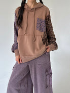 A person wearing a color-blocked hoodie with a solid tan front, patterned brown sleeves, and a purple floral chest pocket, paired with wide-leg mauve cargo pants against a plain light gray wall.