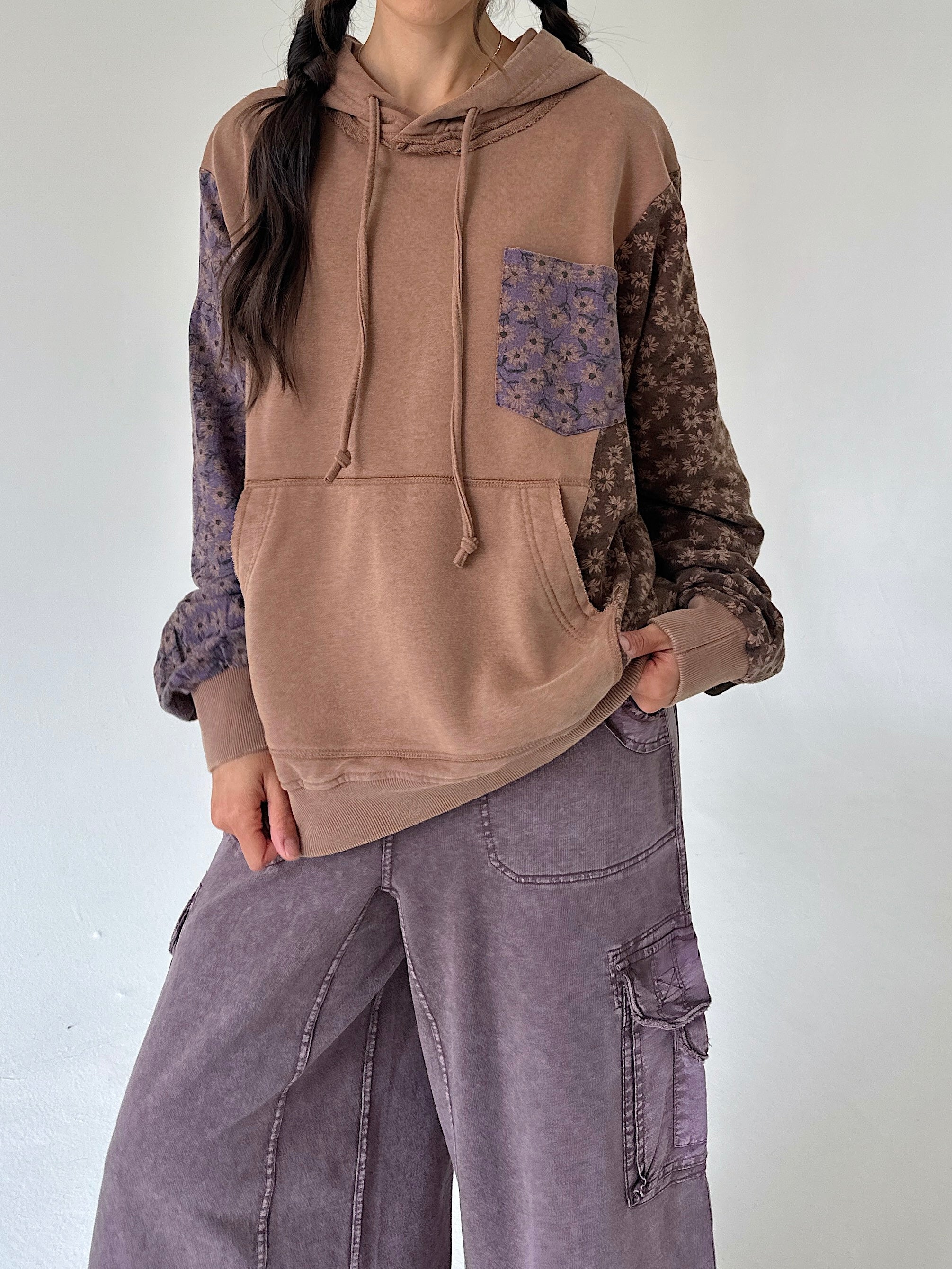 A person wearing a color-blocked hoodie with a solid tan front, patterned brown sleeves, and a purple floral chest pocket, paired with wide-leg mauve cargo pants against a plain light gray wall.