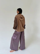 A person standing barefoot in a studio, shown from the back, wearing a brown hooded sweatshirt with patterned sleeves and wide-leg mauve-purple cargo pants.