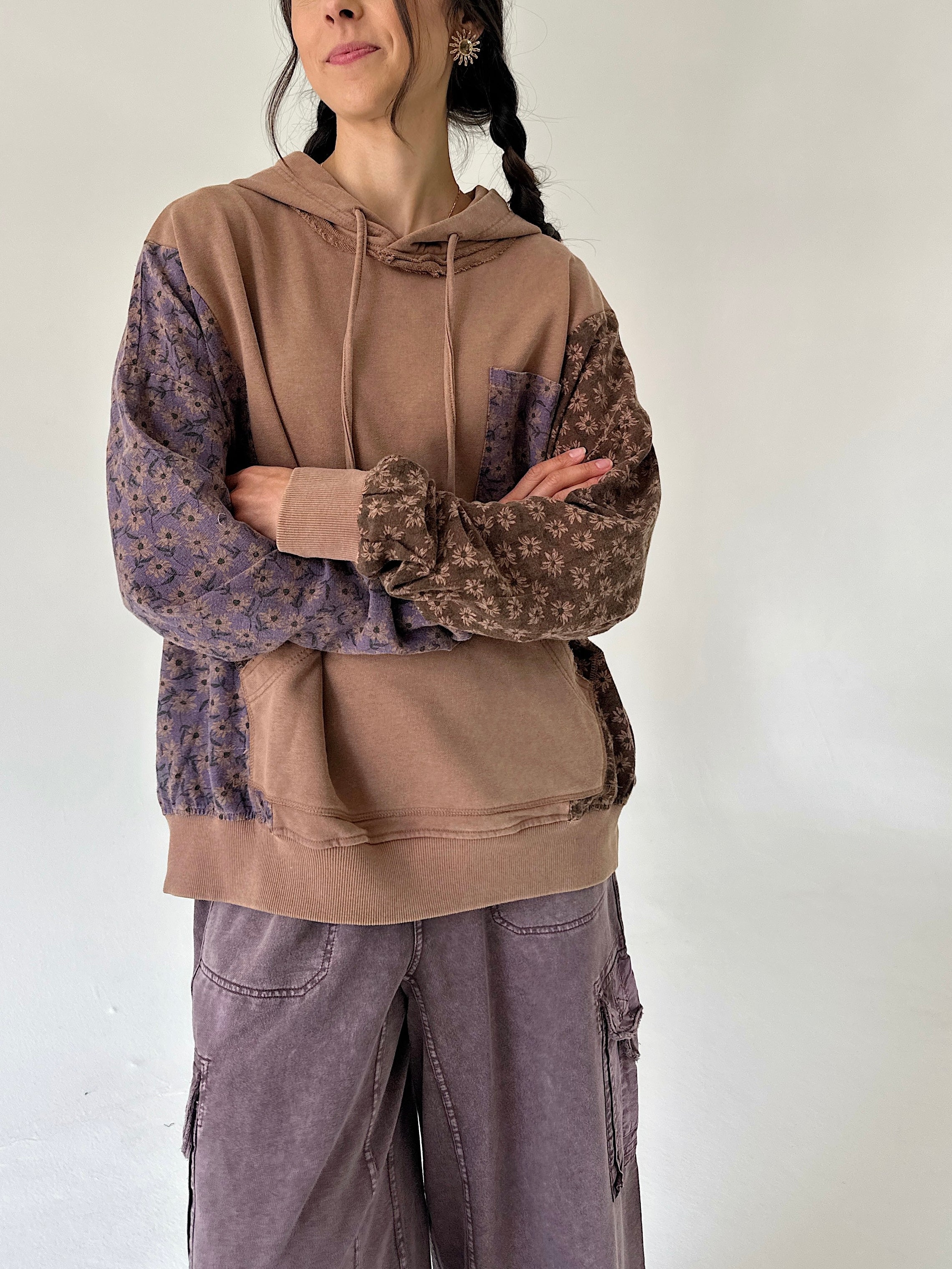 A woman wearing a color-blocked hoodie with a solid tan body and patterned purple sleeves, paired with wide-leg mauve cargo pants, standing against a plain off-white wall.