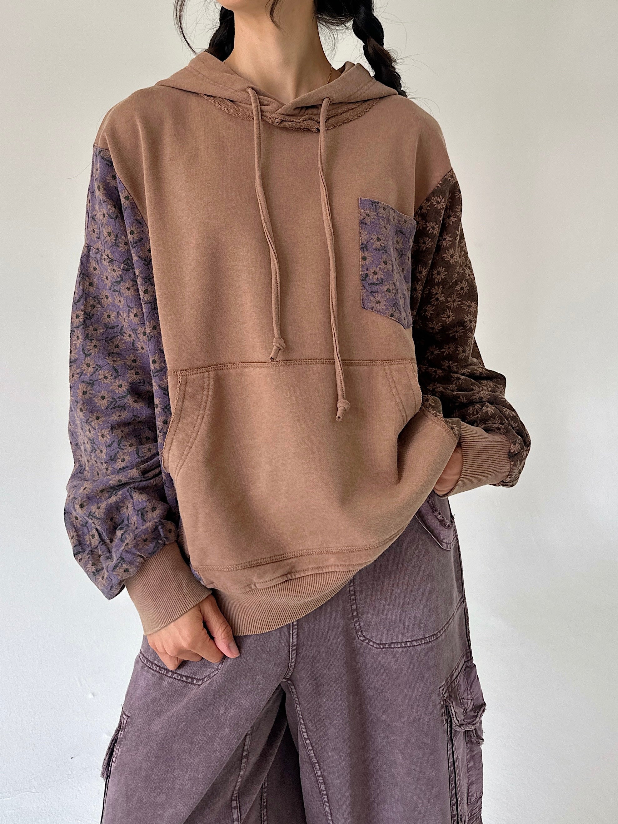 A person wearing a color-block hoodie with a solid tan front, purple floral patterned sleeves, and a dark brown floral chest pocket. The person is also wearing wide-leg mauve cargo pants against a plain off-white wall.