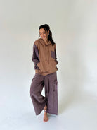 A woman modeling a two-tone hoodie and wide-leg cargo pants set in a minimalist studio setting. The hoodie features a tan front with dark purple textured sleeves and pocket details.