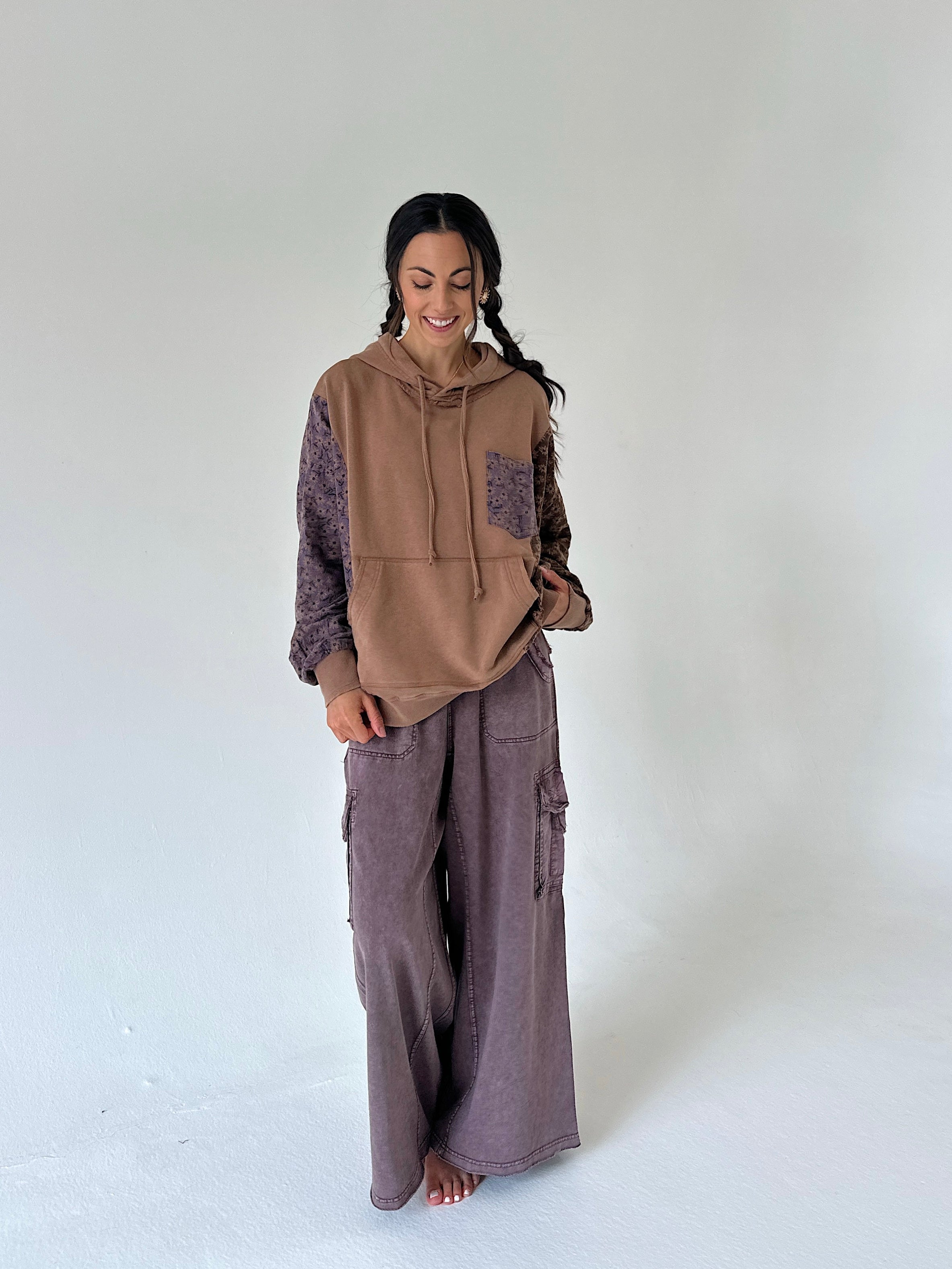 A woman stands barefoot against a plain light gray studio background, wearing a brown and mauve-purple patchwork hoodie with matching wide-leg cargo pants.