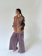 A woman modeling a brown and purple patchwork hoodie with patterned sleeves and wide-leg mauve-purple cargo pants, standing barefoot against a plain white studio background.