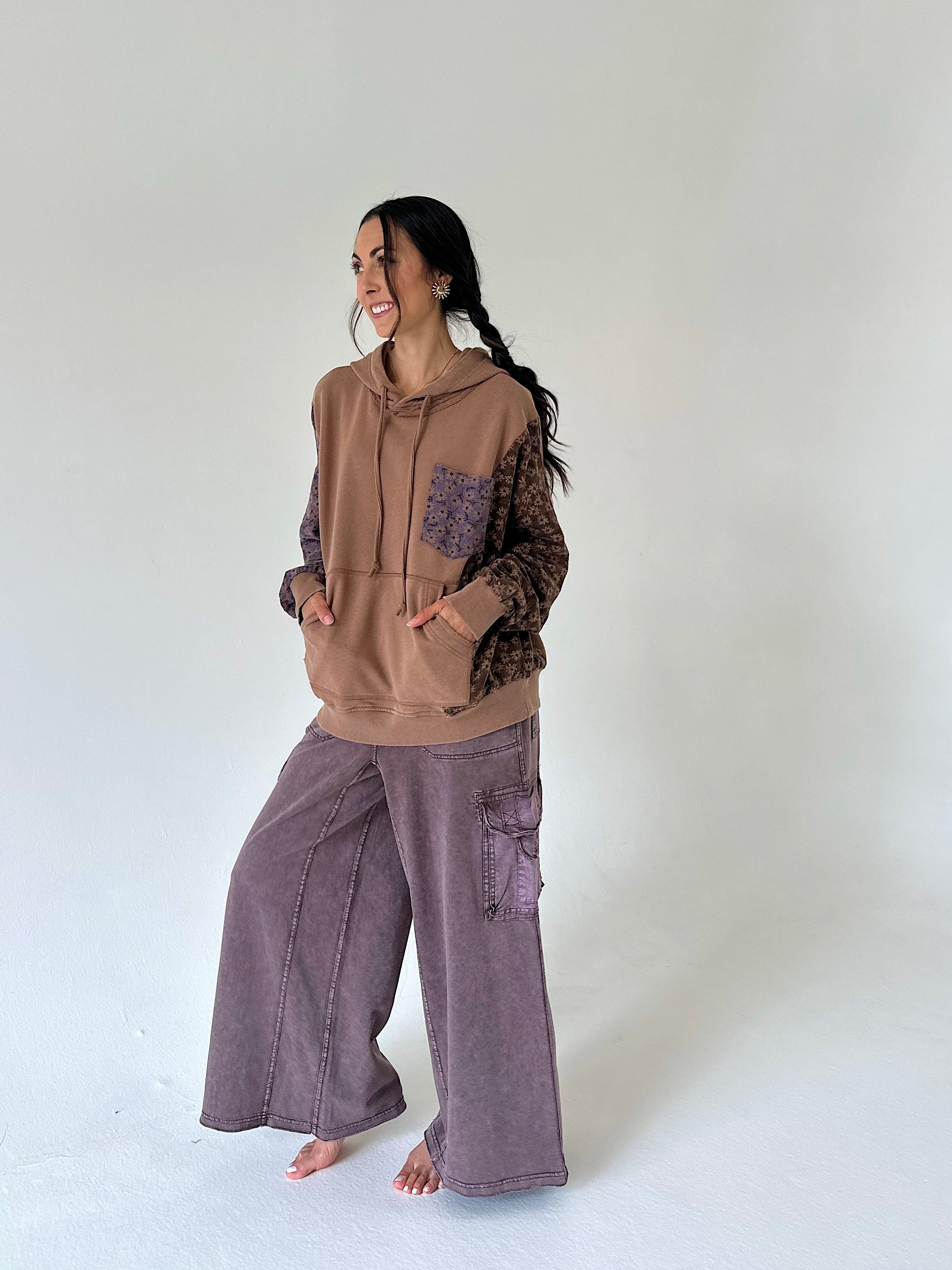 A woman modeling a brown and purple patchwork hoodie with patterned sleeves and wide-leg mauve-purple cargo pants, standing barefoot against a plain white studio background.