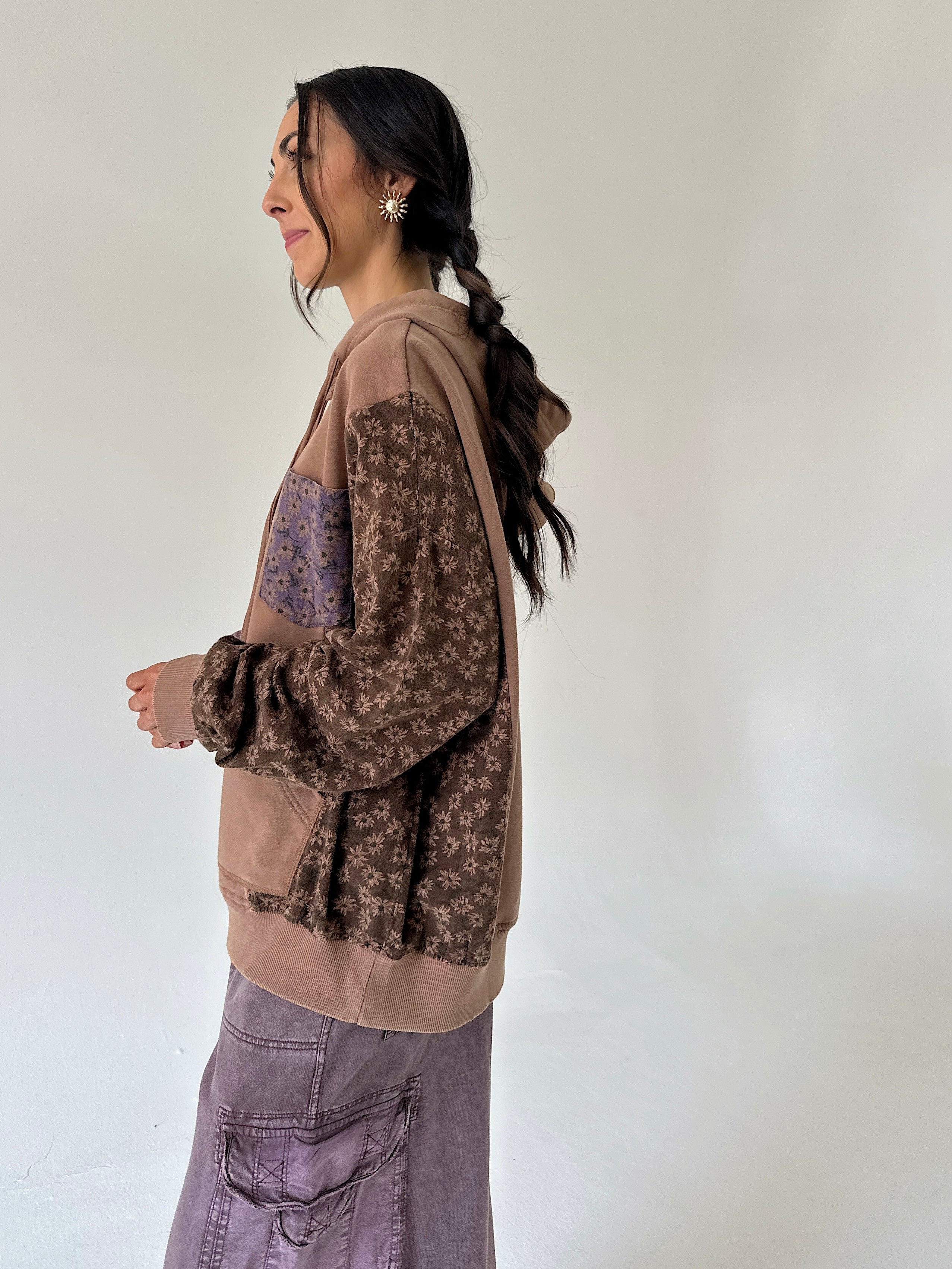 A woman shown in profile wearing a patchwork hoodie with brown floral sleeves and a solid tan body, paired with a purple skirt, against a plain off-white background.