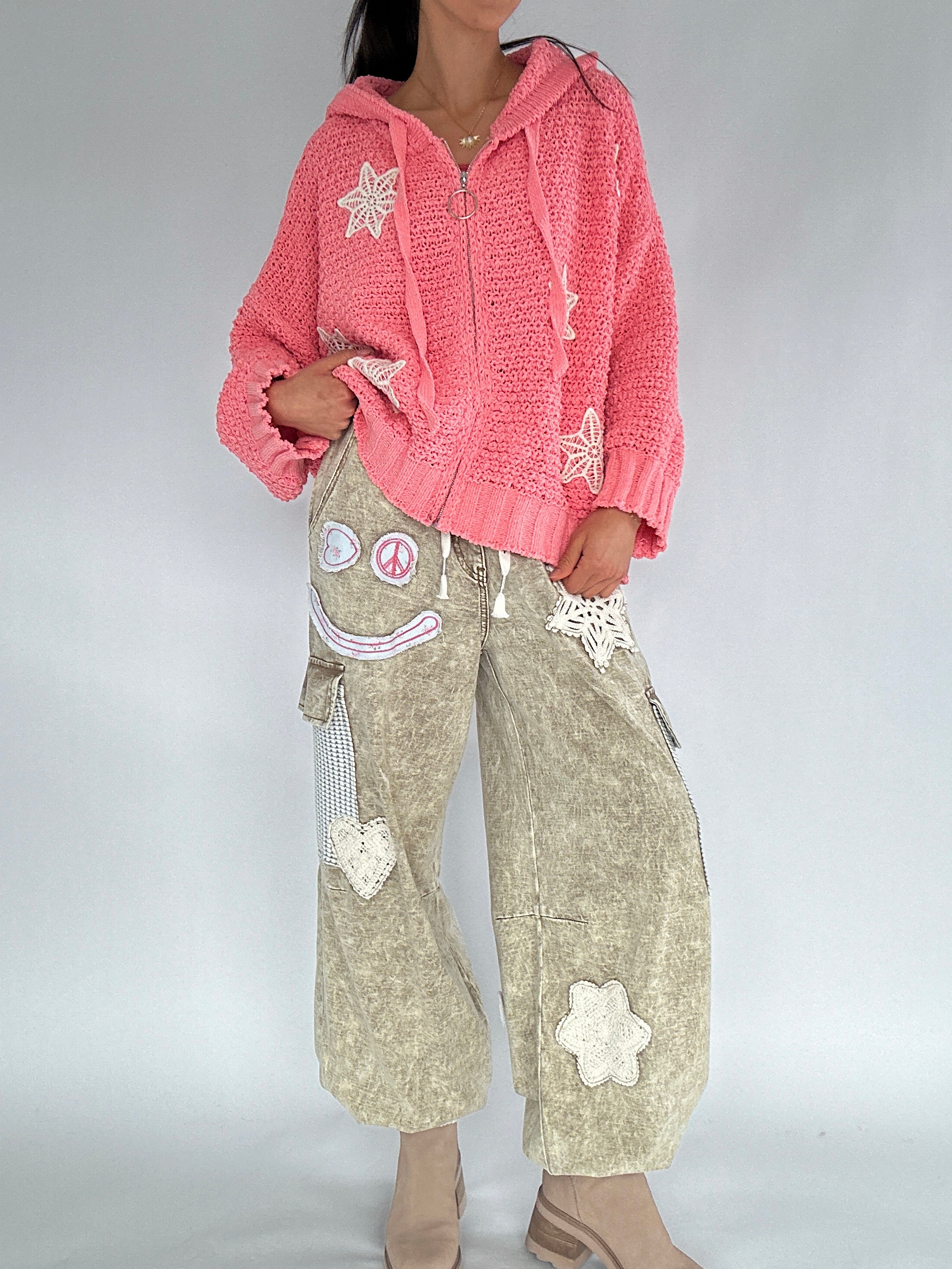 Person wearing a pink knitted hoodie and beige pants with floral patches on a plain background