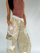 Person wearing a brown tank top and beige pants with decorative patches on a plain background