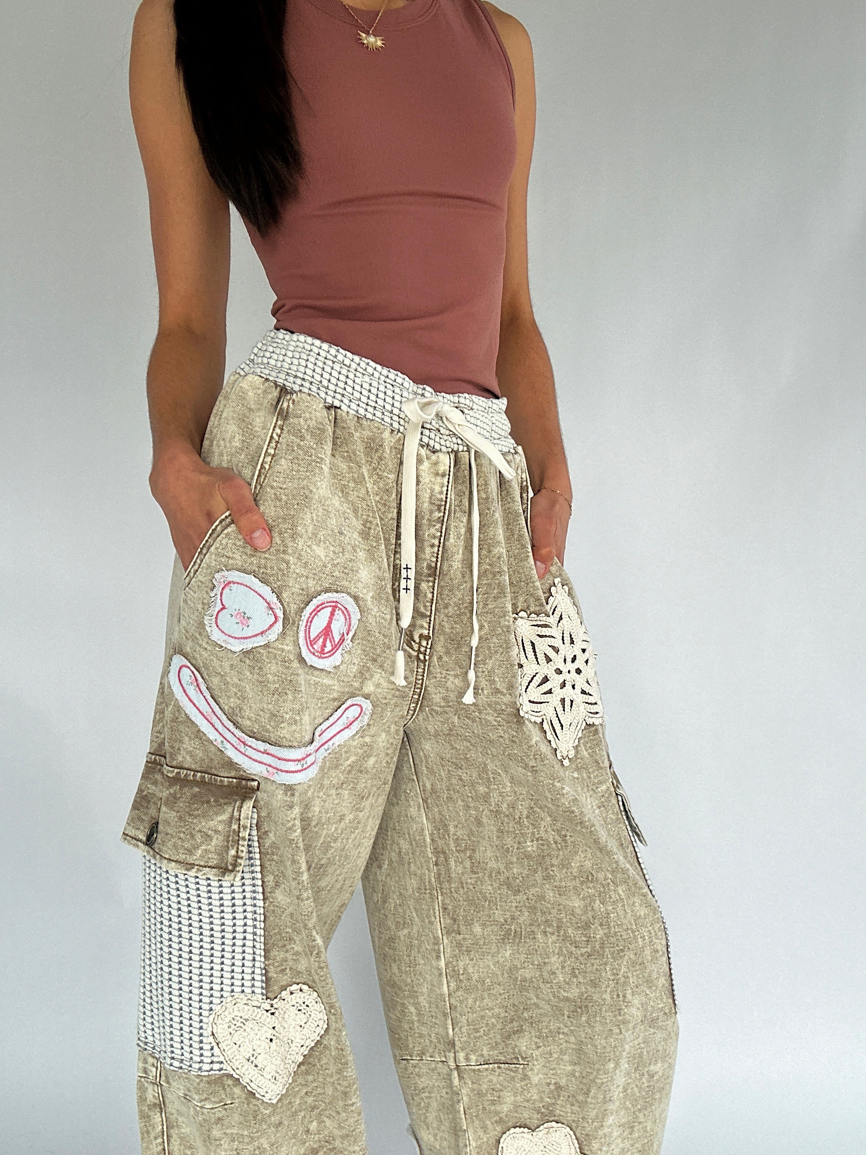 Person wearing a brown tank top and beige pants with decorative patches on a plain background