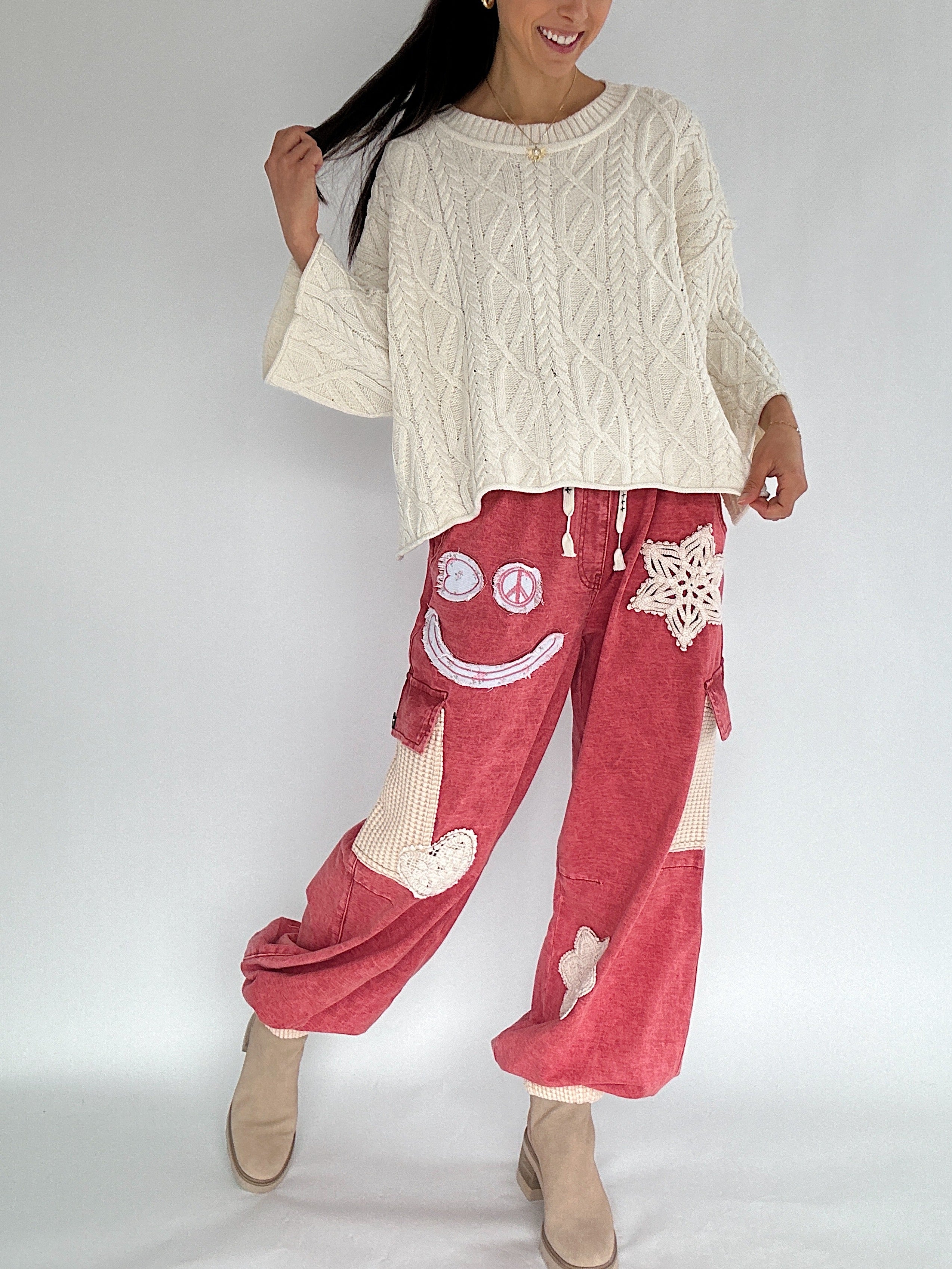 Person wearing a cream cable knit sweater and red pants with smiley face designs on a white background