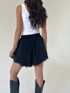 A woman shown from the back wearing a white sleeveless top, black ruffled shorts, and dark cowboy boots against a plain light background.